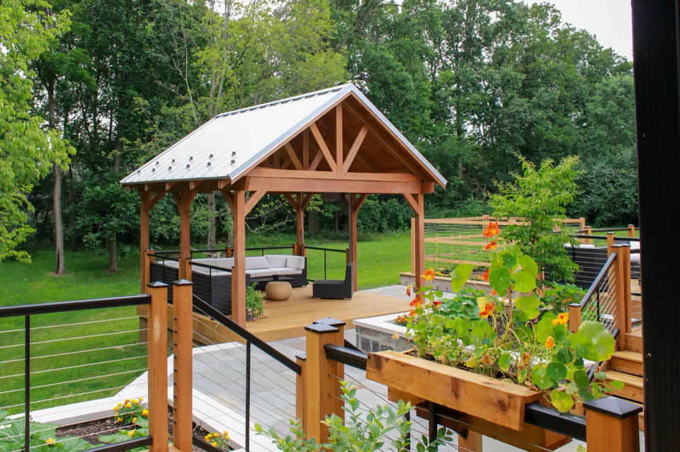 Covered wood pavilion viewed from deck with railing, planter box, lawn, and wooded backdrop.
